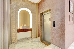 Un ascensor moderno con detalles metálicos y un panel de control, junto a un suelo de baldosas de mármol y una pared con un diseño geométrico.
