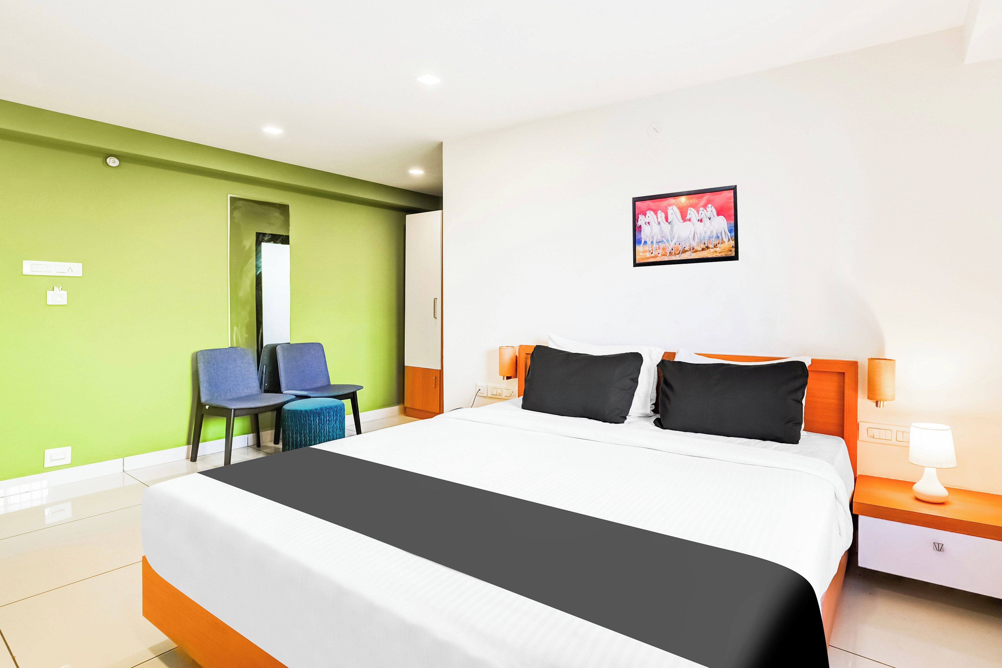 A hotel room with a bed, a desk, two chairs, a painting, and a nightstand with a lamp.