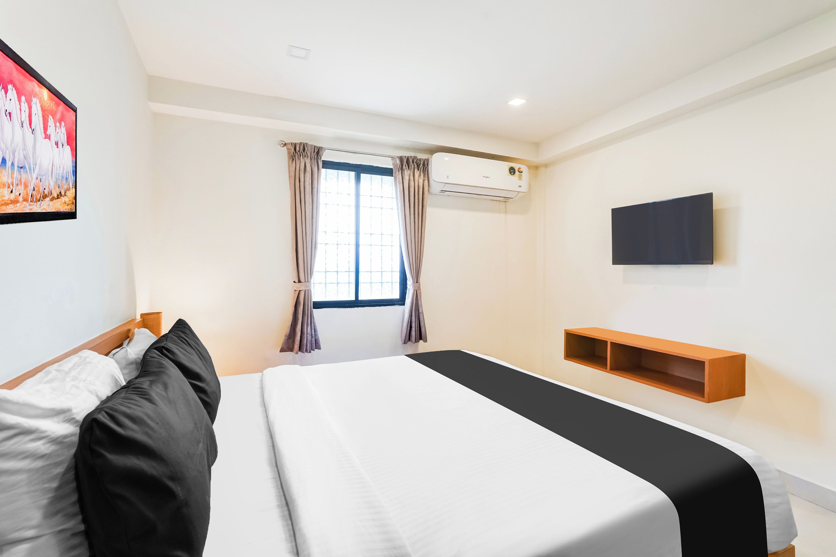 A hotel room with a bed, a flat-screen TV, an air conditioning unit, a window with curtains, and a wall-mounted shelf.