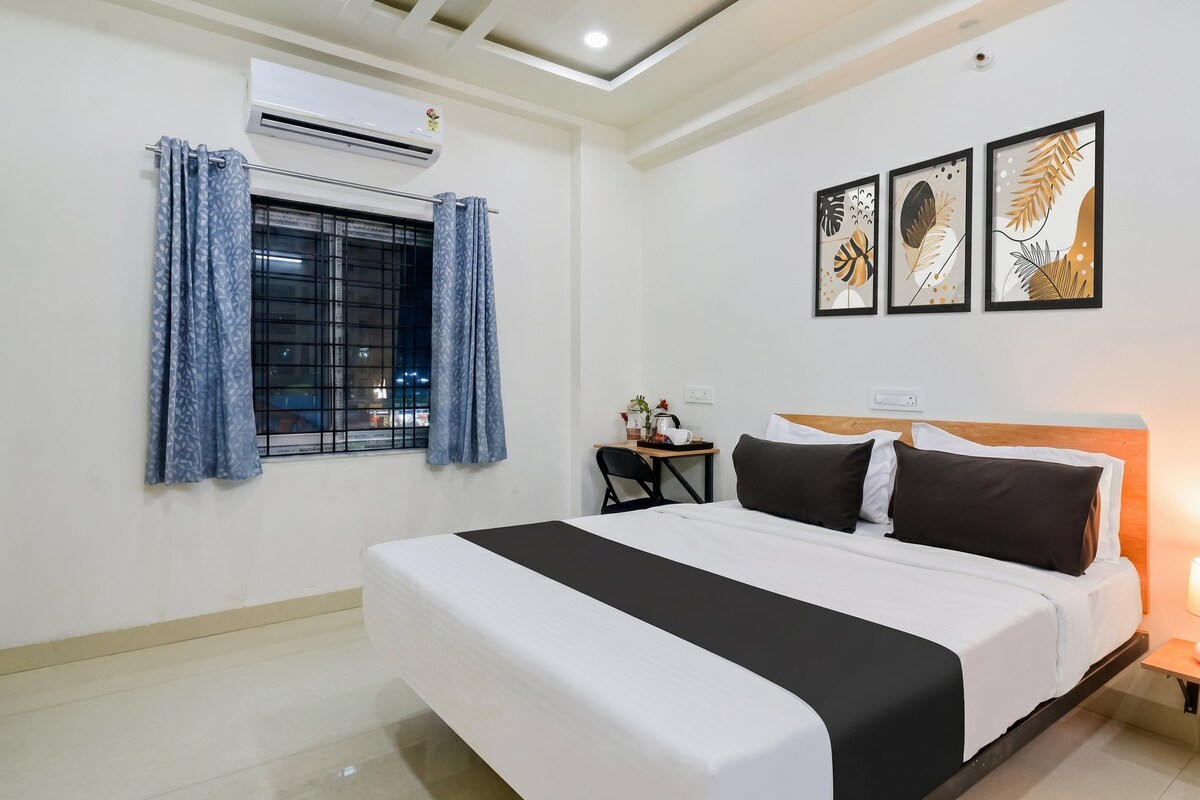 A hotel room with a large bed, a desk with a chair, an air conditioning unit, and a window with curtains.