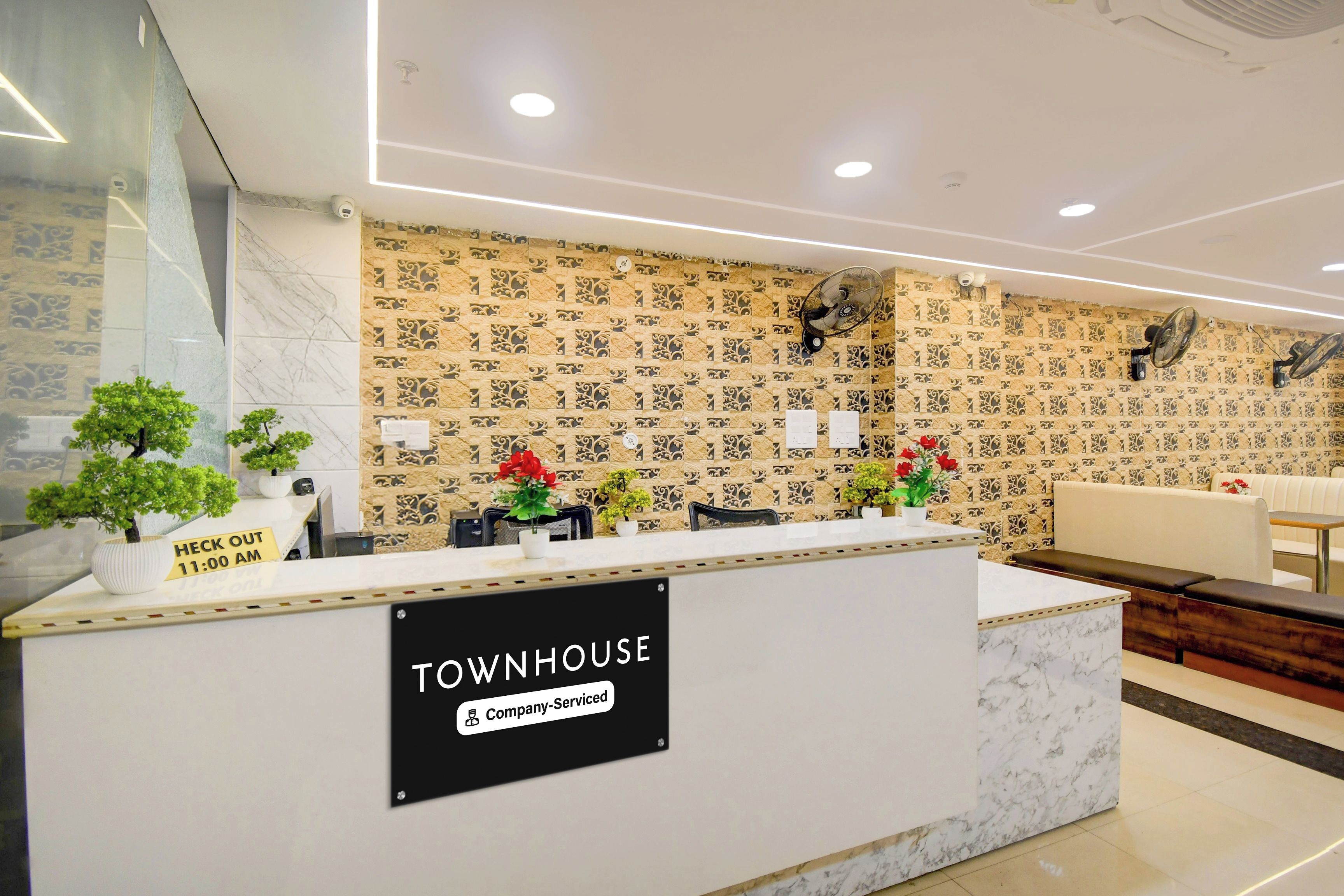 Una mesa de recepción con un letrero que dice 'Townhouse Company-Serviced'. Hay plantas en macetas y un reloj que marca 'Fin de la estancia 11:00 AM'.
