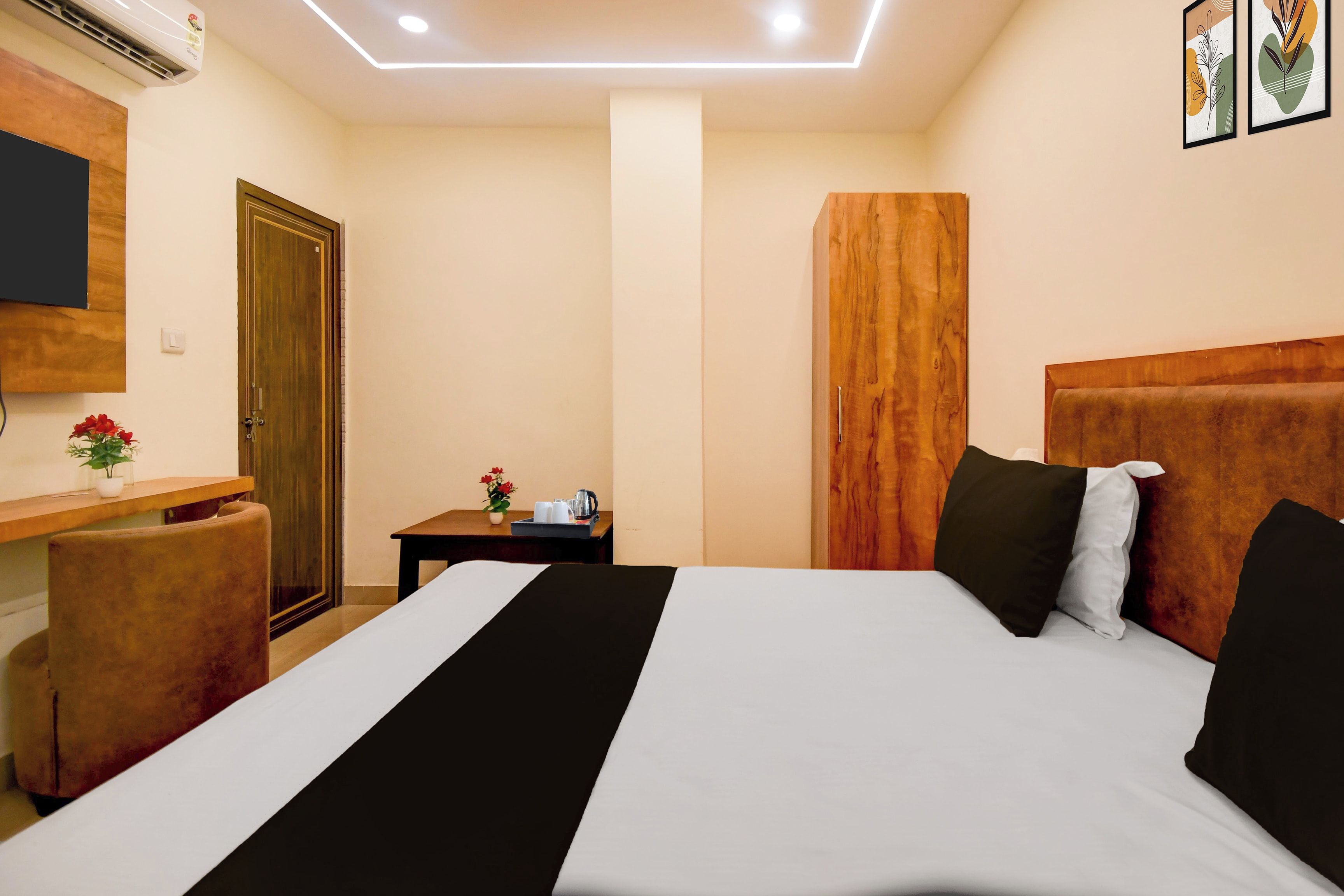 Habitación de hotel con cama, mesita de noche, una pequeña mesa con un florero y un televisor montado en la pared, además de un cabecero de madera.