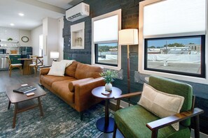 Ein Wohnzimmer mit braunem Sofa, einem grünen Sessel, einem kleinen Tisch und einer Lampe. Im Hintergrund ist eine Küche mit einem Kühlschrank und einer Wanduhr zu sehen.