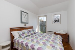 Ein Schlafzimmer mit einem hölzernen Bett, einem kleinen weißen Nachttisch, zwei gerahmten Kunstwerken an der Wand und einem Korbwäschekorb mit Handtüchern.