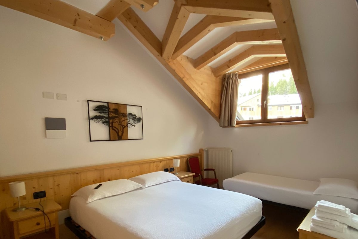 A room with a wooden ceiling, a bed, a small table with towels, a chair, and a window.