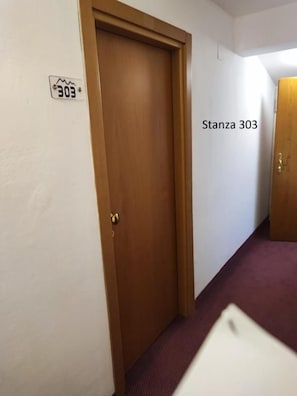 Un pasillo con una puerta de madera cerrada, una pared blanca con un cartel con número de puerta y suelo alfombrado.