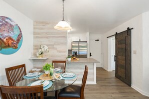 A dining area with a glass table, wooden chairs, and a pendant light. - 1BD Garden View Retreat with AC Near South Maui Beaches- Kauhale Makai 108 (Kihei)