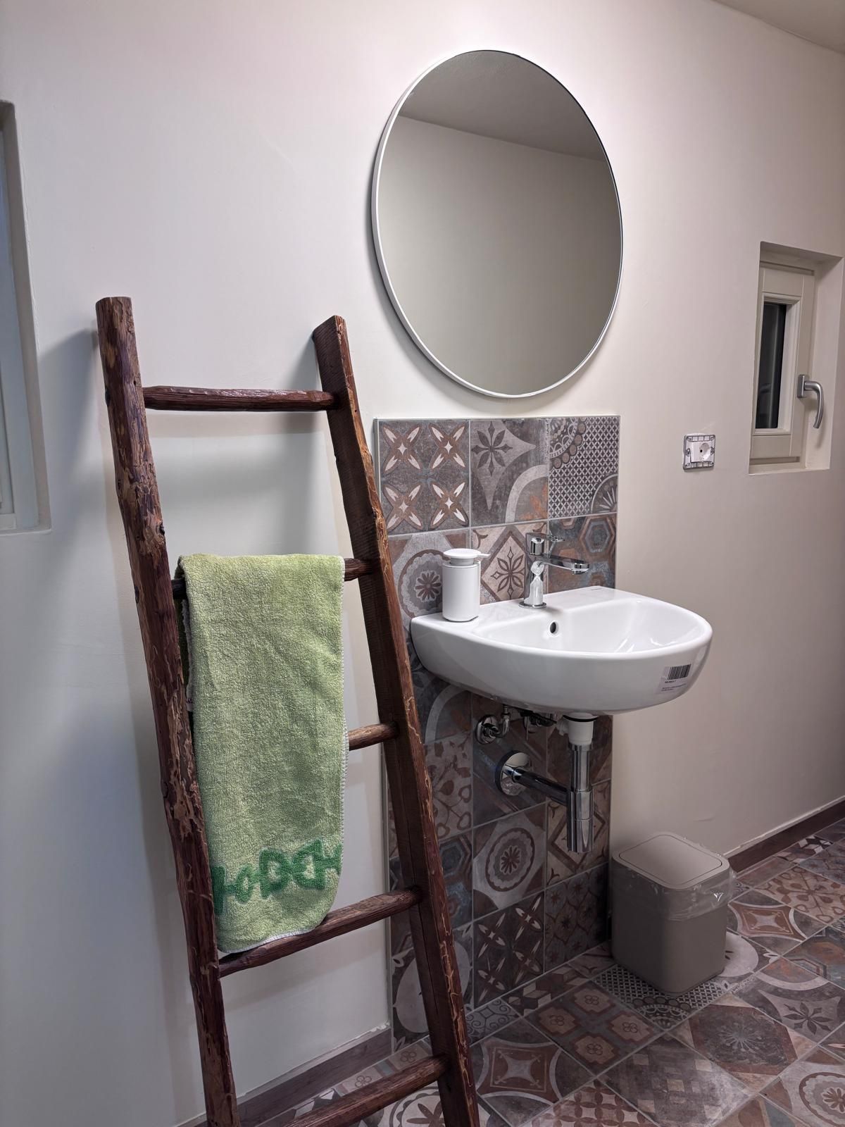 A bathroom with a wooden ladder, a white sink, a round mirror, and patterned tiles.