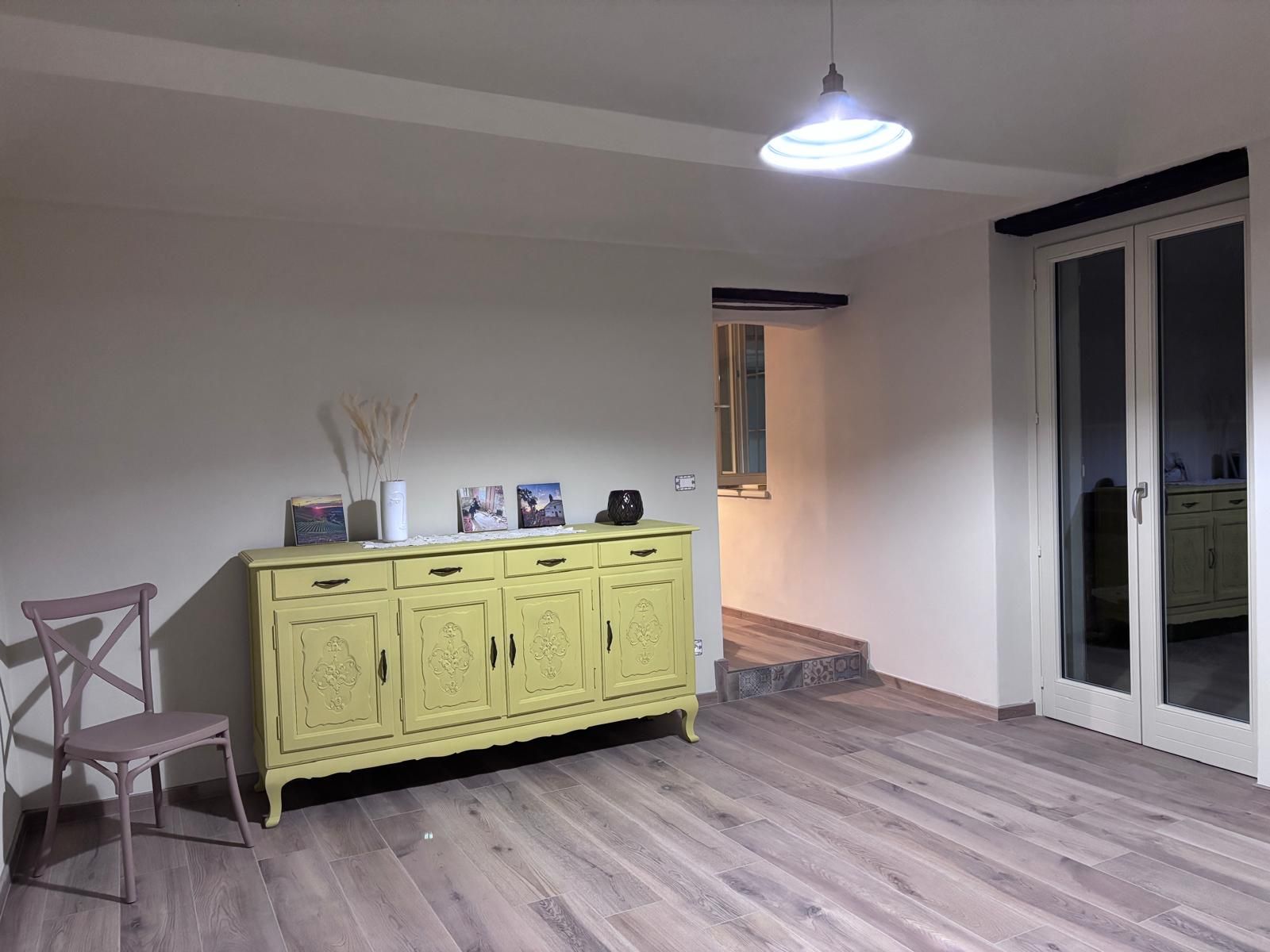 Quarto com piso de madeira, um armário amarelo com detalhes entalhados, uma cadeira e luminária no teto.