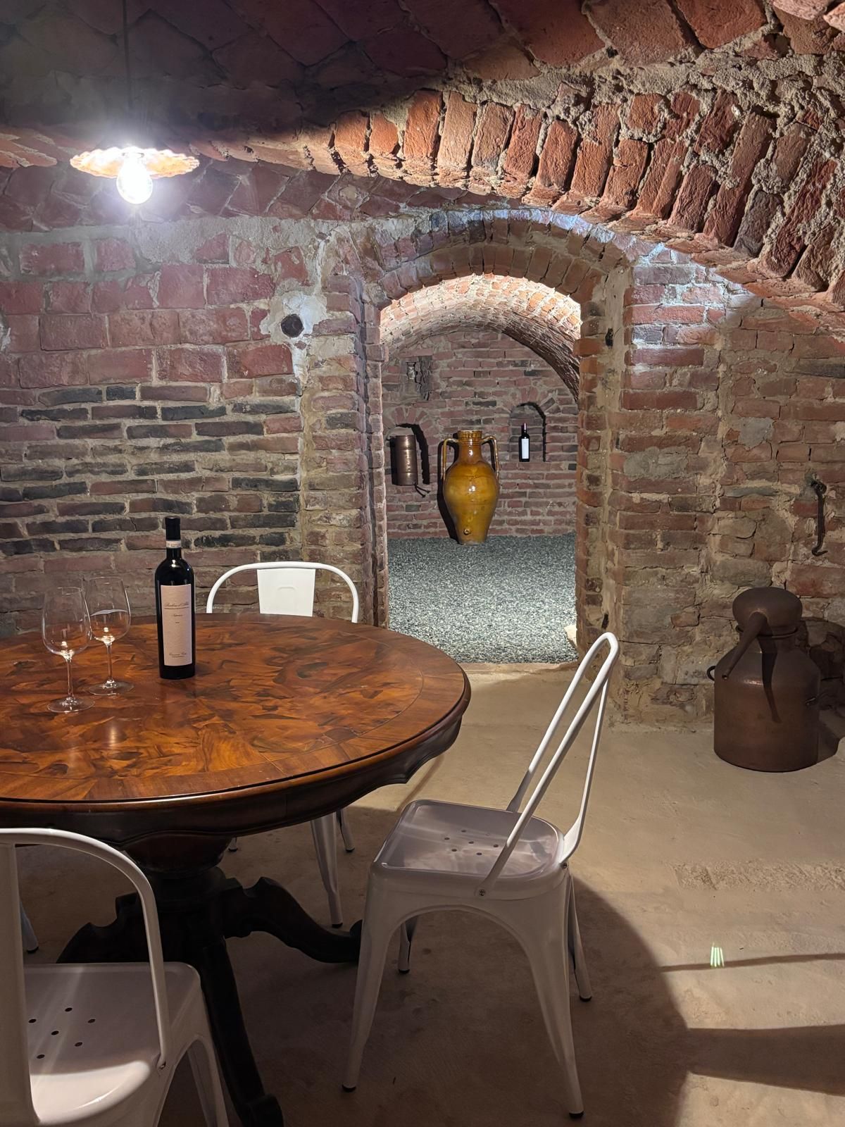 Un coin repas rustique avec un mur en briques, une table en bois, des chaises blanches, une bouteille de vin et un grand vase.