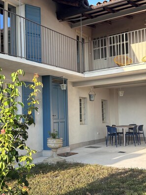 Un edificio a due piani con un balcone, una zona pranzo con sedie e una pianta in vaso.