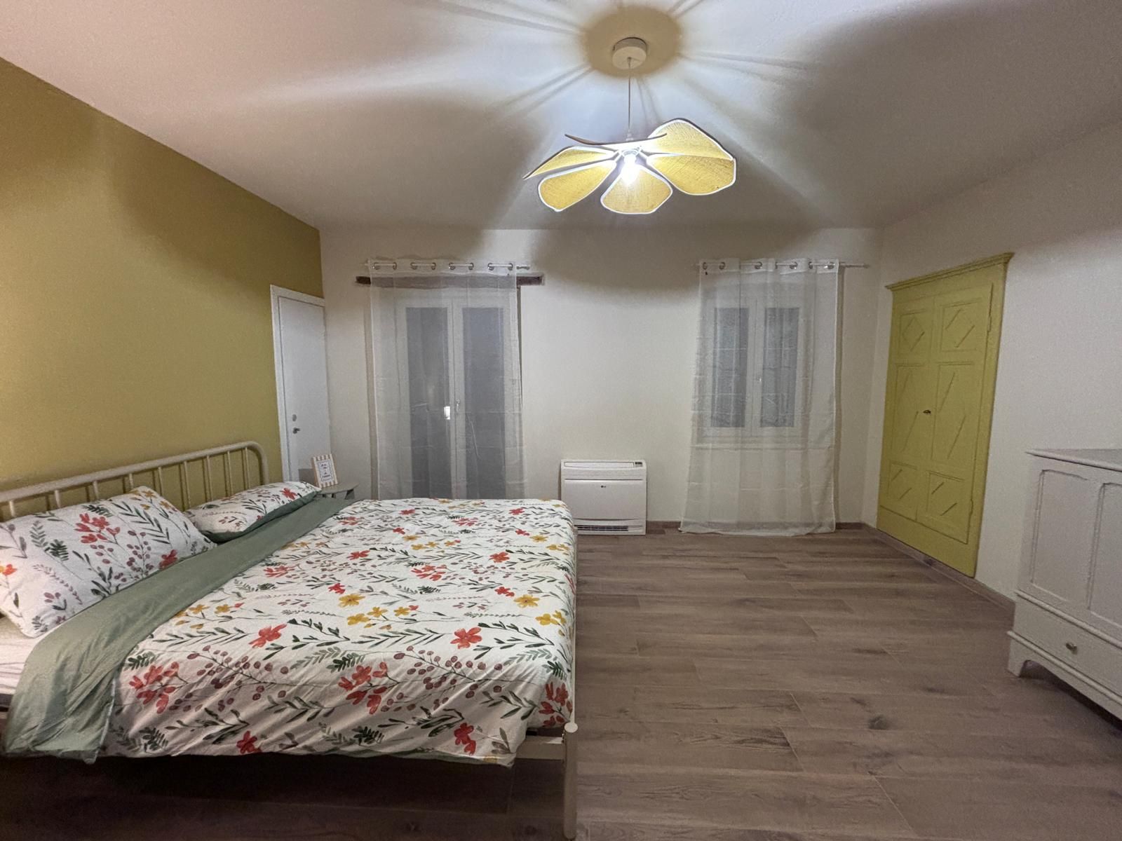 Une chambre à coucher avec un lit, un ventilateur de plafond, une fenêtre avec des rideaux et une armoire.