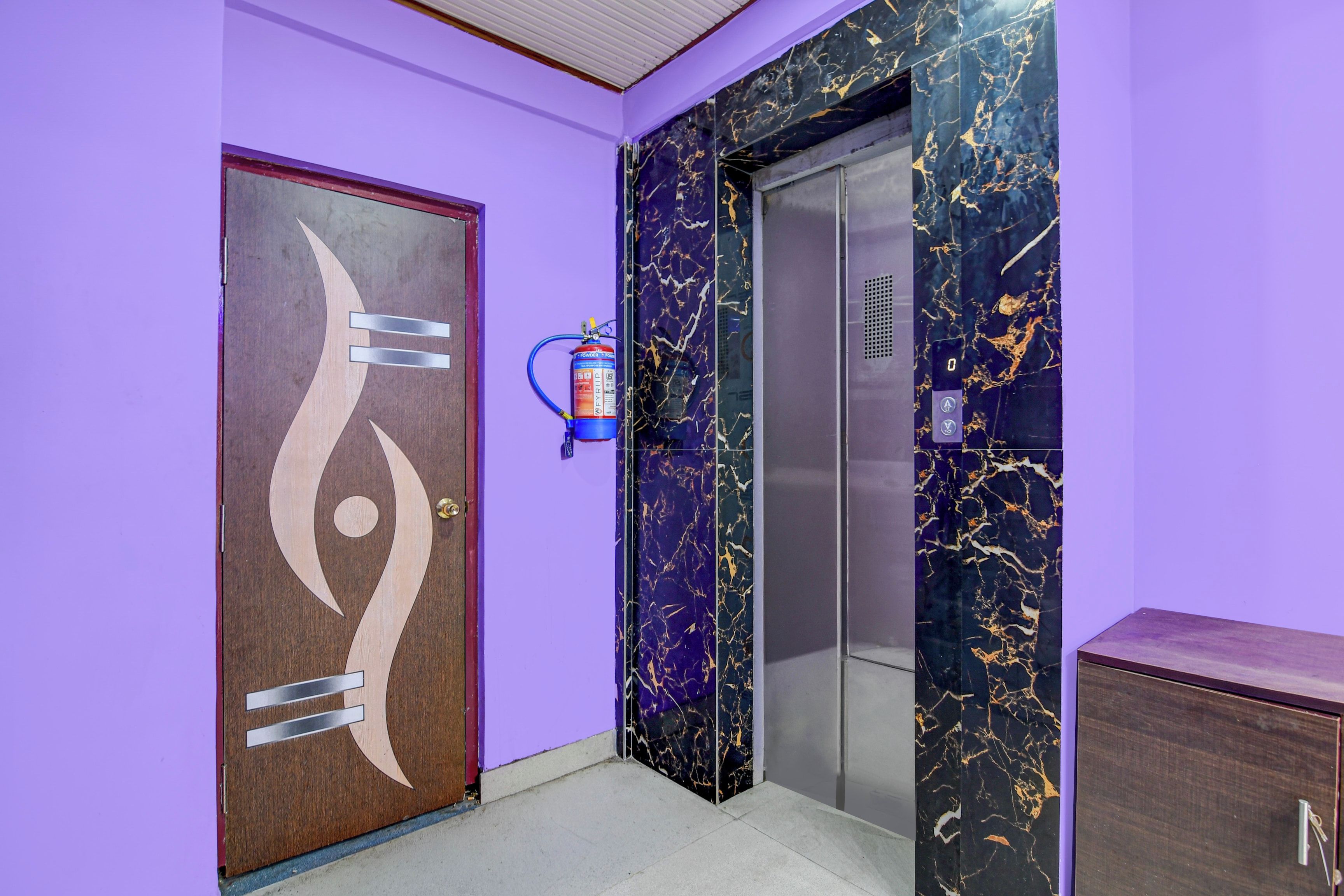 A hallway with a purple wall, a wooden door with a unique design, and an elevator with a marble finish.