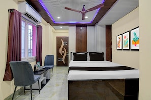 Habitación de hotel con cama, escritorio, silla, unidad de aire acondicionado, ventilador de techo y una obra de arte en la pared.