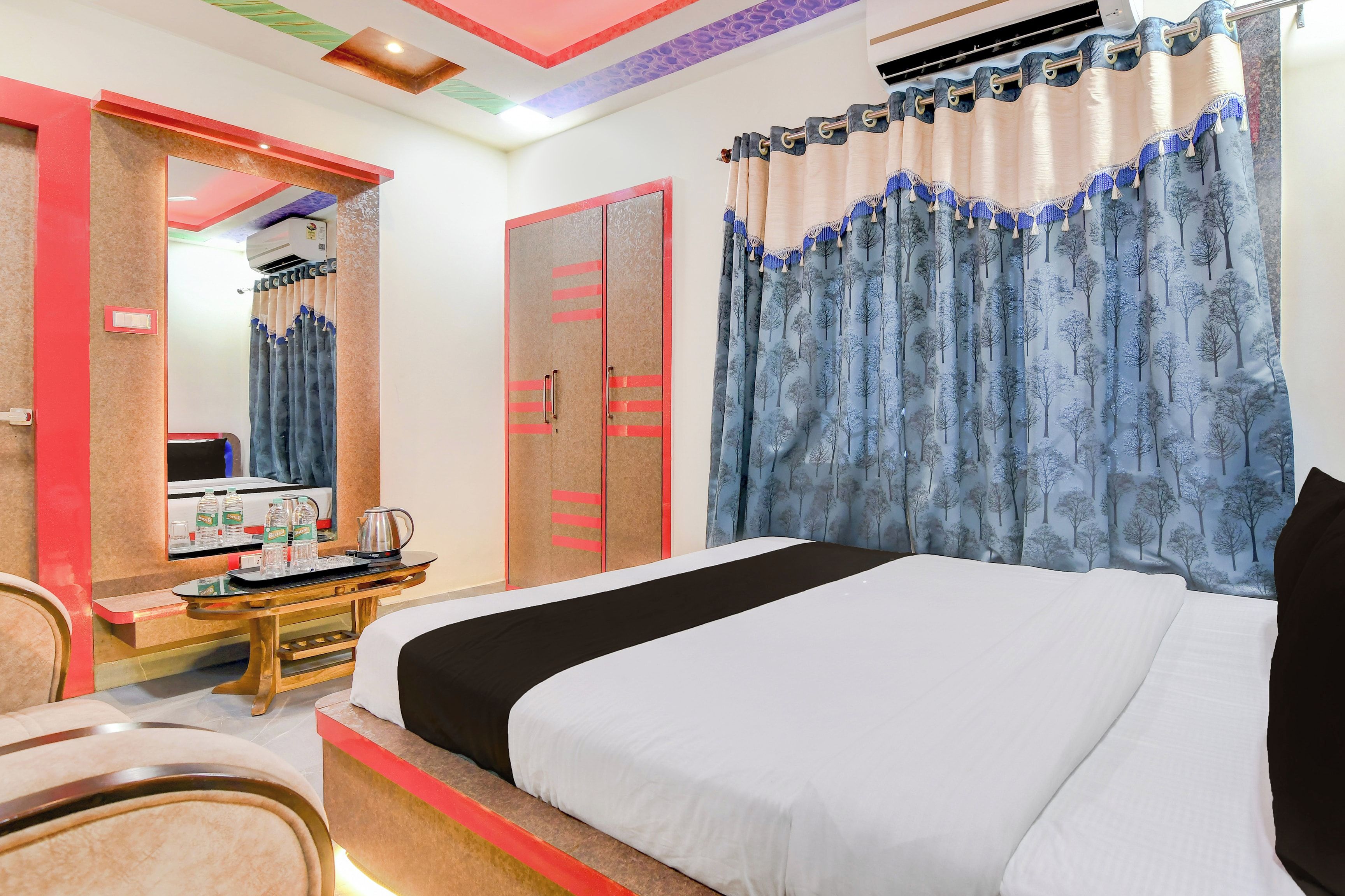 A hotel room with a bed, a desk with a kettle, a mirror, and a wardrobe.