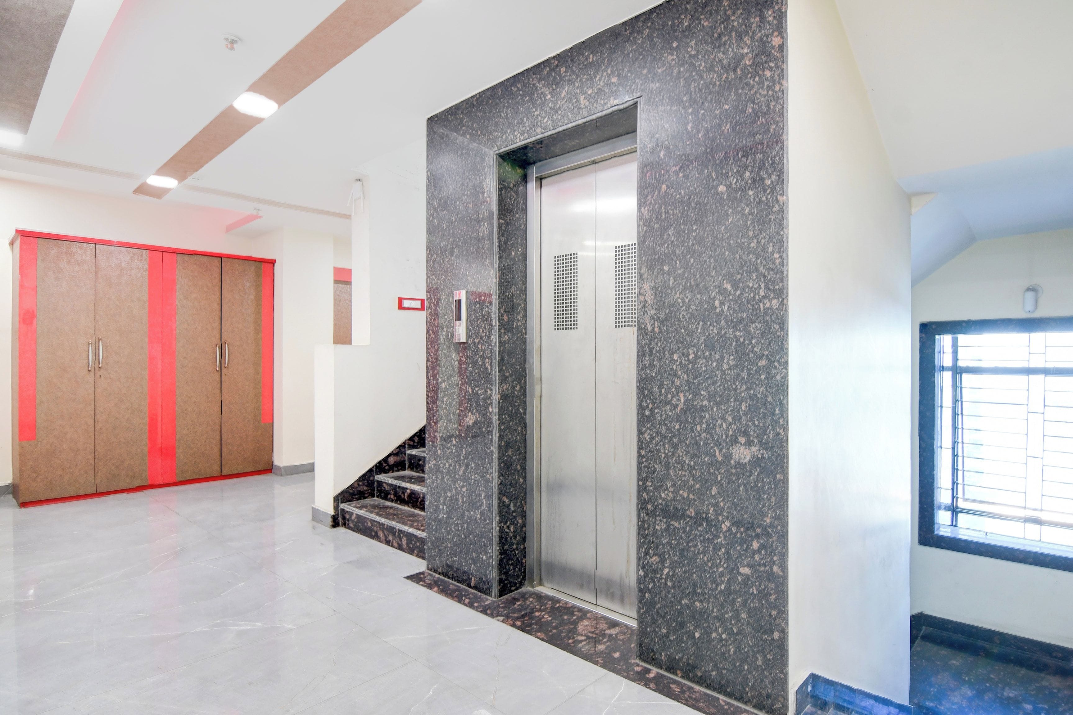 A modern elevator with a granite facade, a red-framed door, and a staircase leading to an upper floor.
