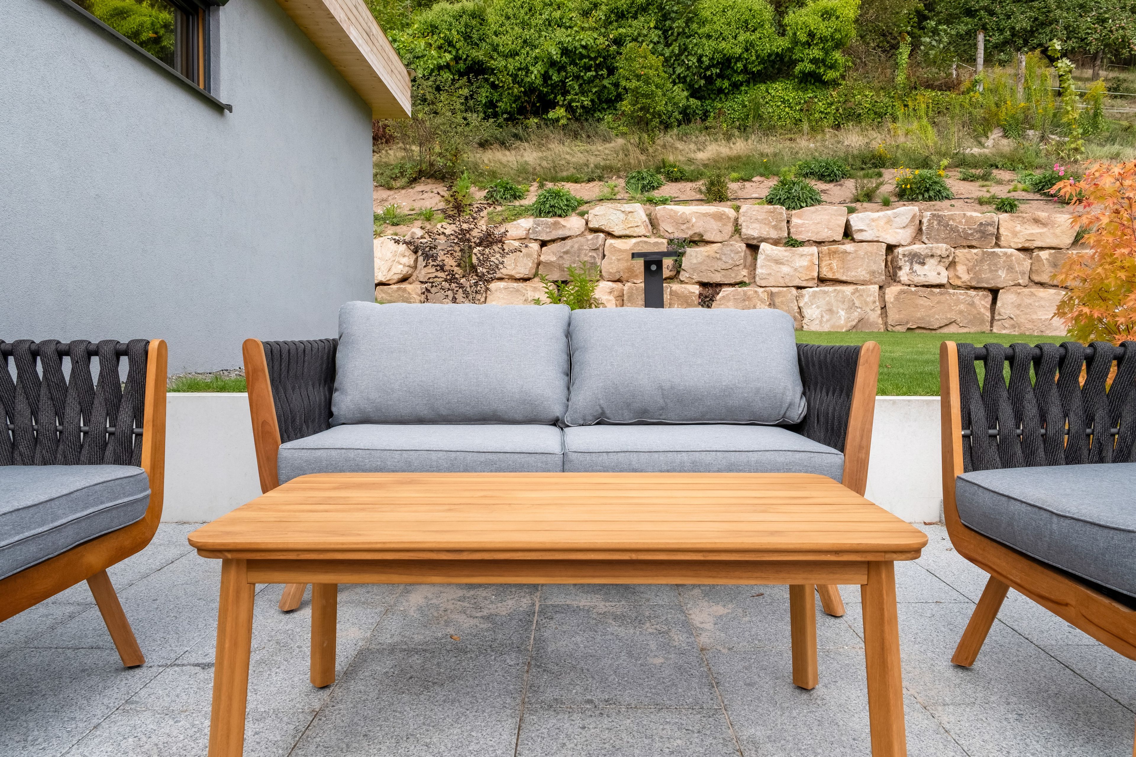 Eine Terrasse mit einem grauen Sofa, zwei Holzstühlen und einem Holztisch.