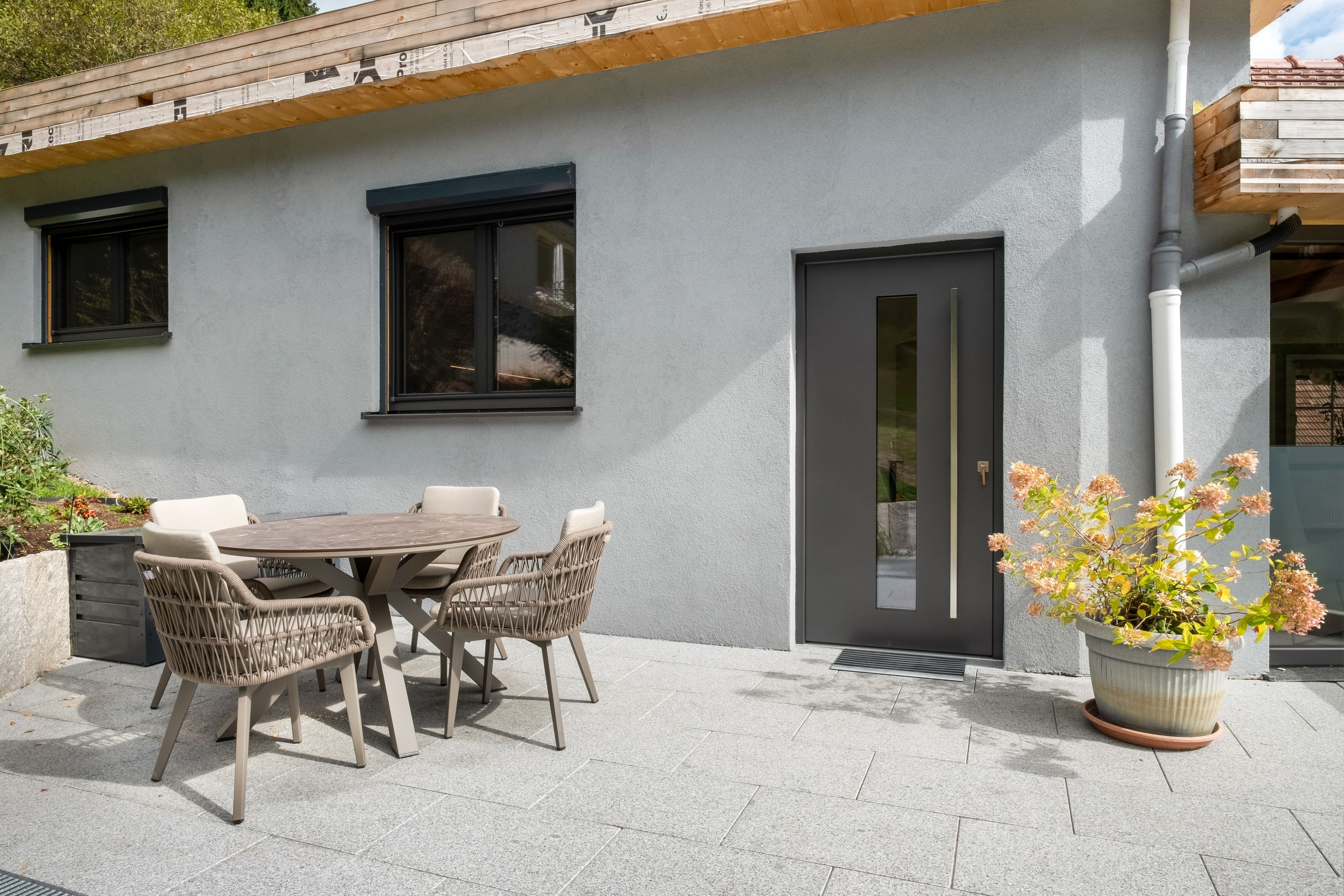 A modern outdoor patio with a round table and wicker chairs, a grey wall, and a door with a vertical handle.
