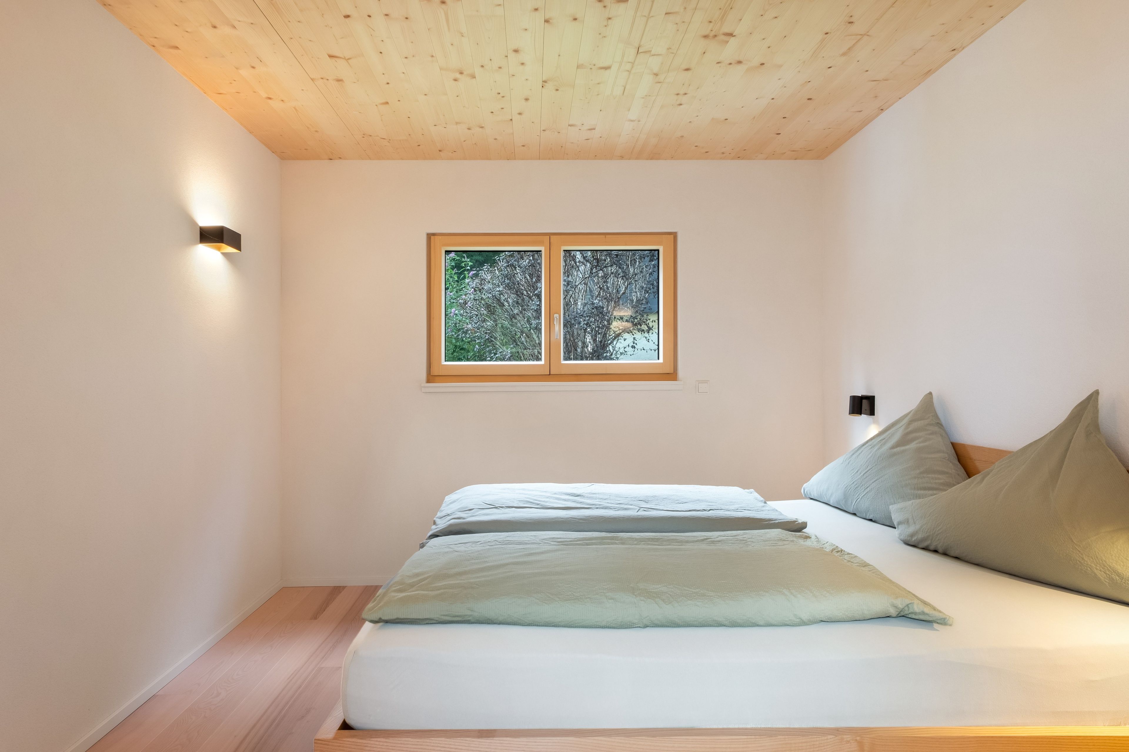 A bedroom with a wooden ceiling, a bed with pillows, a nightstand, and a window with a view of trees.