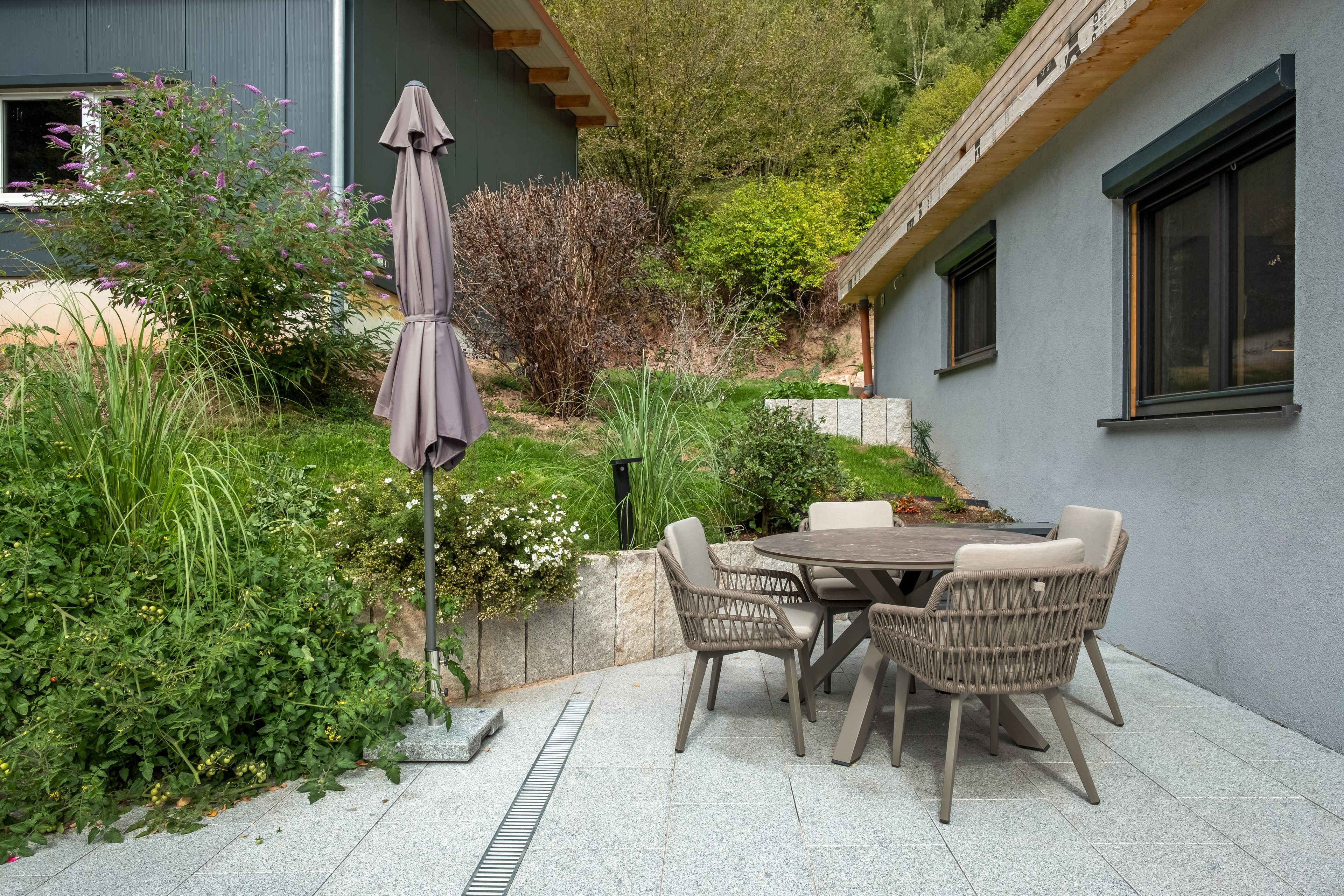 Een terras buiten met een tafel en stoelen, omgeven door groen en een gebouw.