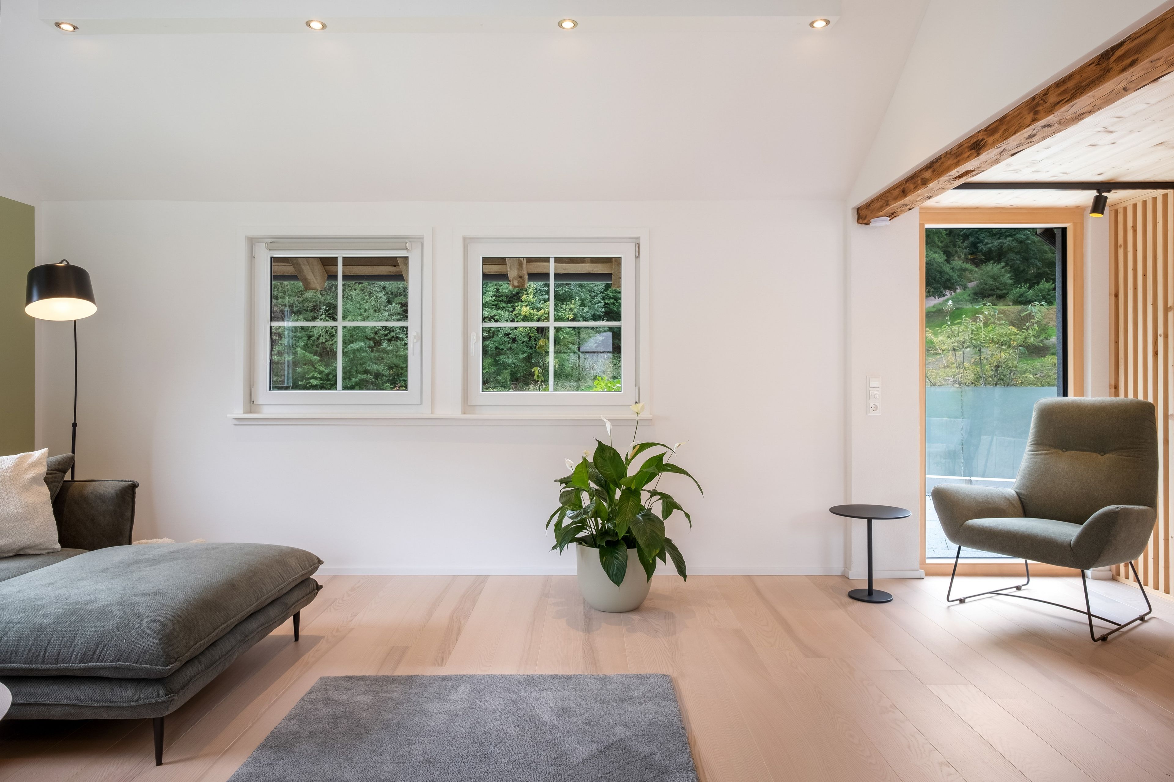 A modern room with a bed, a chair, a floor lamp, and two windows with a view of greenery.
