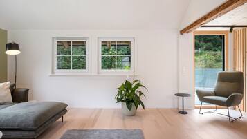 A modern room with a bed, a chair, a floor lamp, and two windows with a view of greenery.