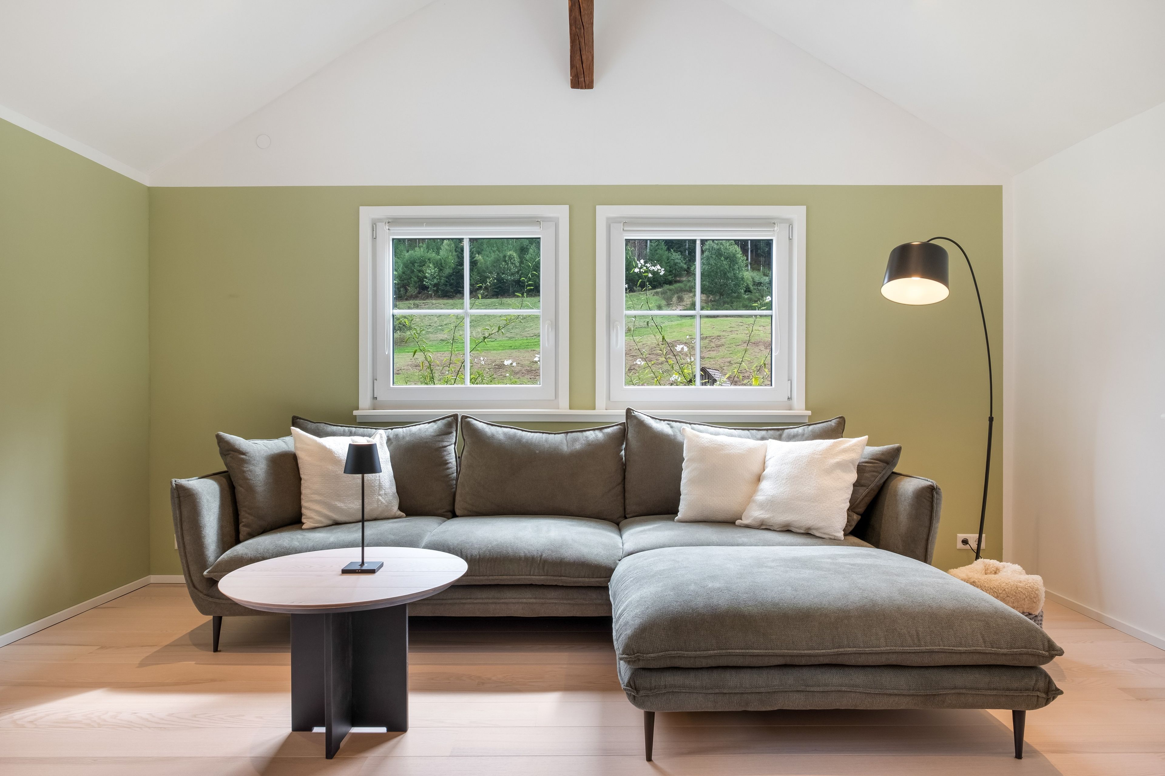 A modern living room with a sectional sofa, a round coffee table, and a floor lamp. There are two windows providing a view of a green landscape.