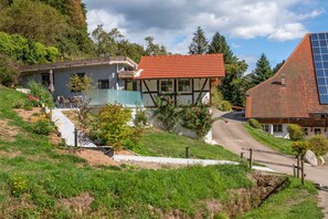 A traditional chalet with a red-tiled roof, surrounded by lush greenery and a clear sky.