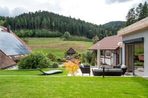 A modern house with a solar panel roof, a patio with outdoor furniture, and a view of a green valley and forested hills.