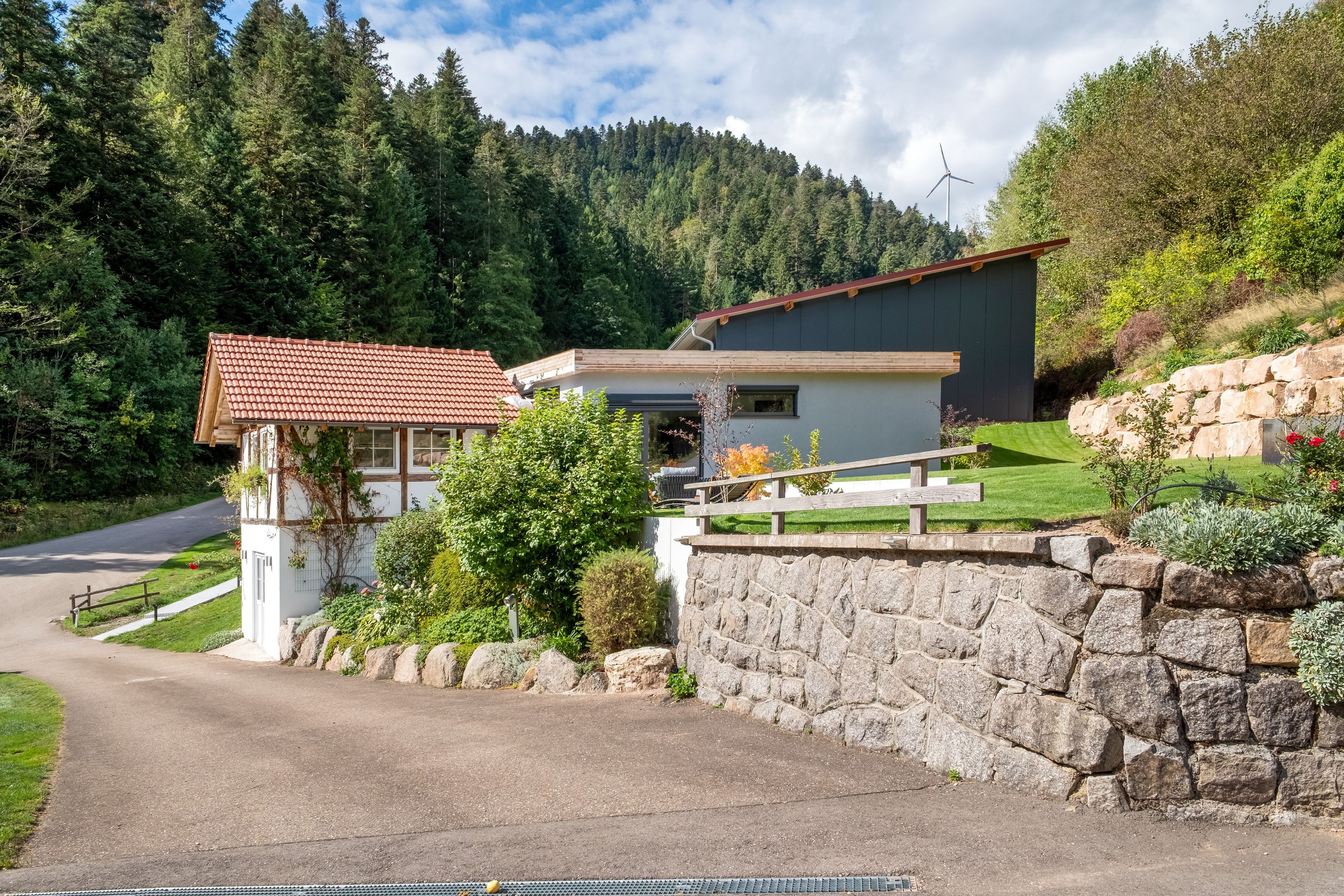 A modern two-story house with a stone retaining wall, a paved driveway, and a forested hillside in the background.