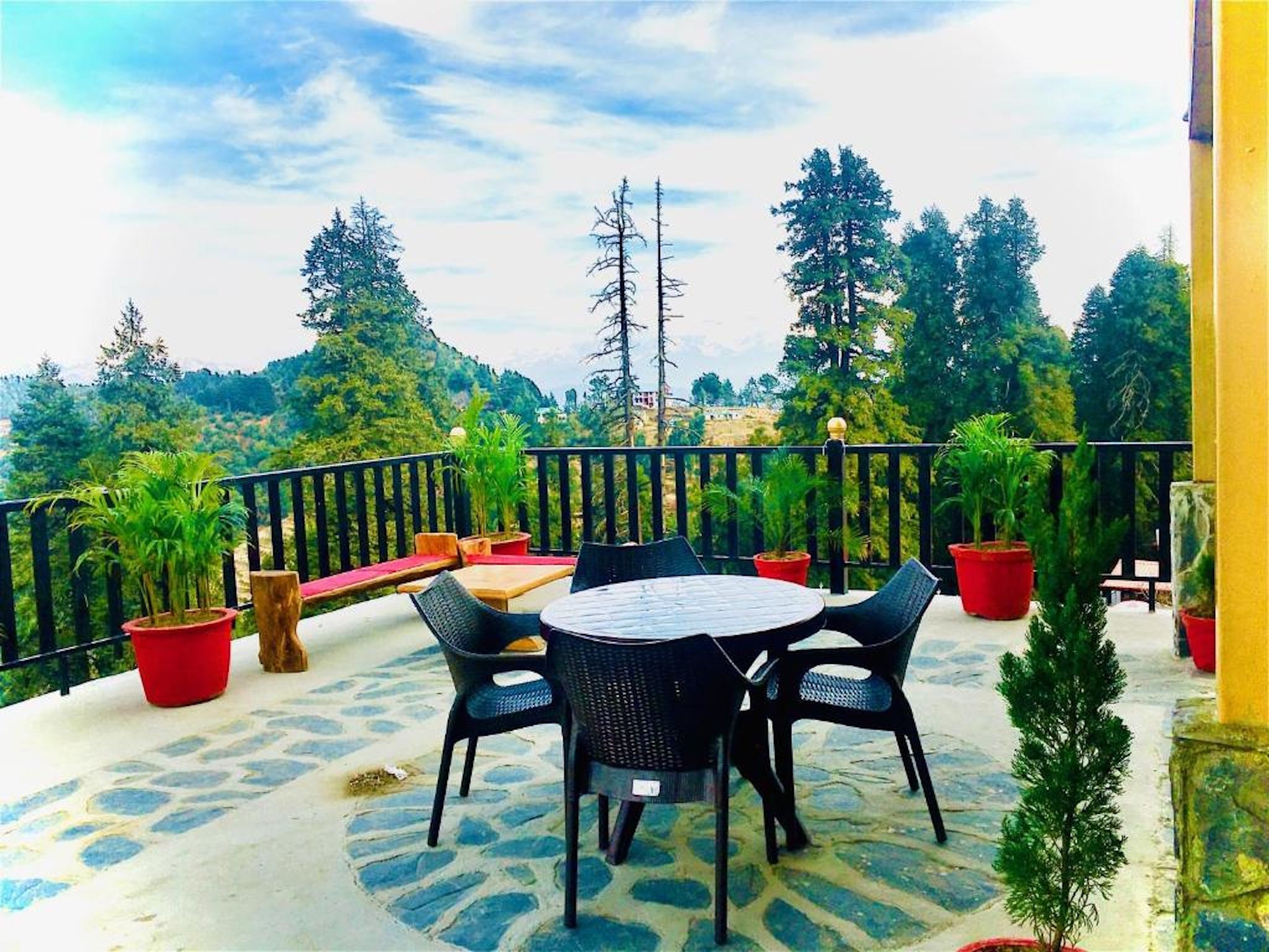 An outdoor patio with a round table and chairs, surrounded by potted plants and a view of a forested hillside.