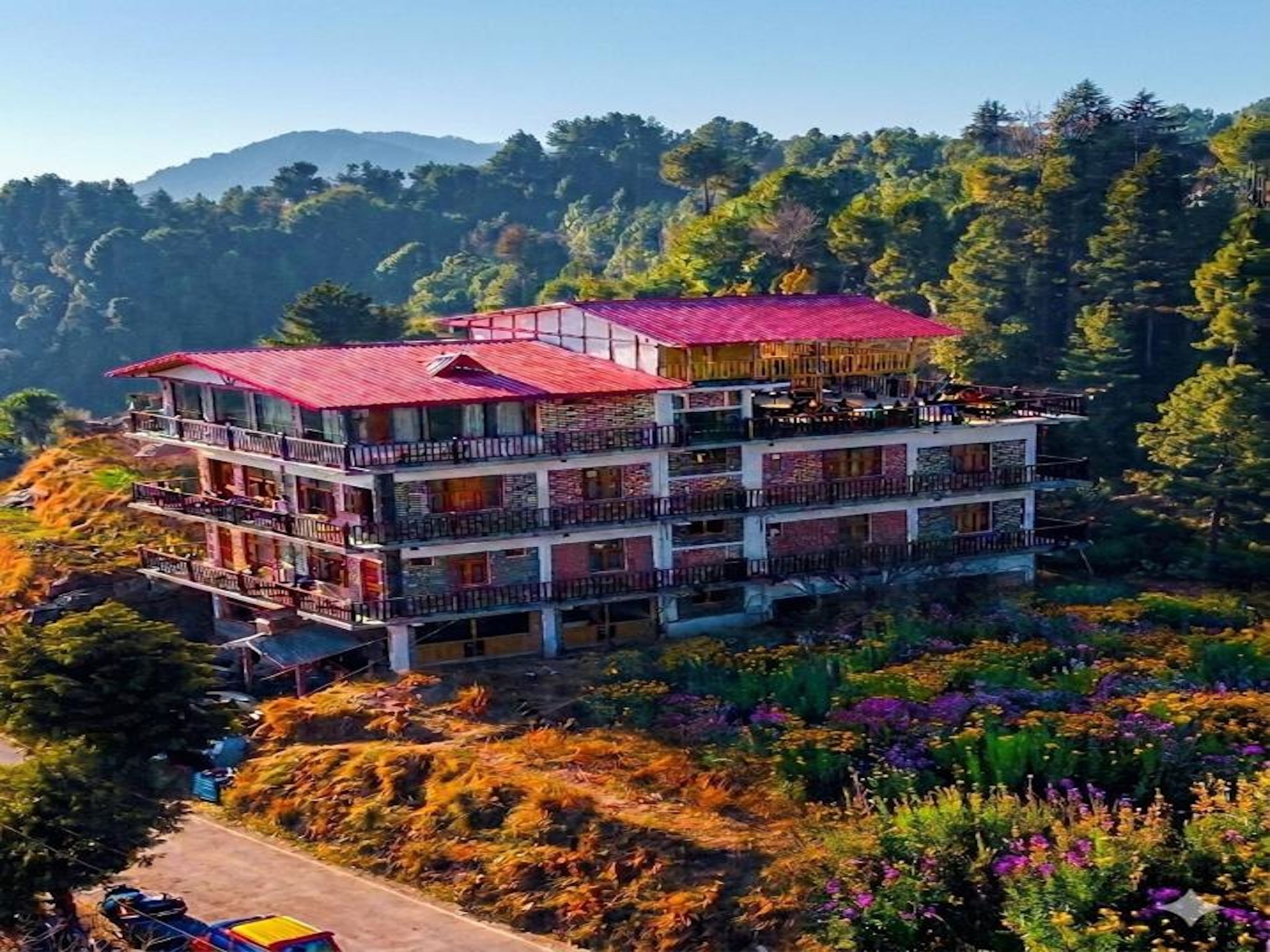 A multi-story hotel with balconies and a red roof, surrounded by lush greenery and mountains.