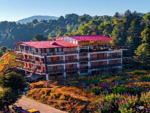 A multi-story hotel with balconies and a red roof, surrounded by lush greenery and mountains. - LA Riqueza Kanatal (Tehri)