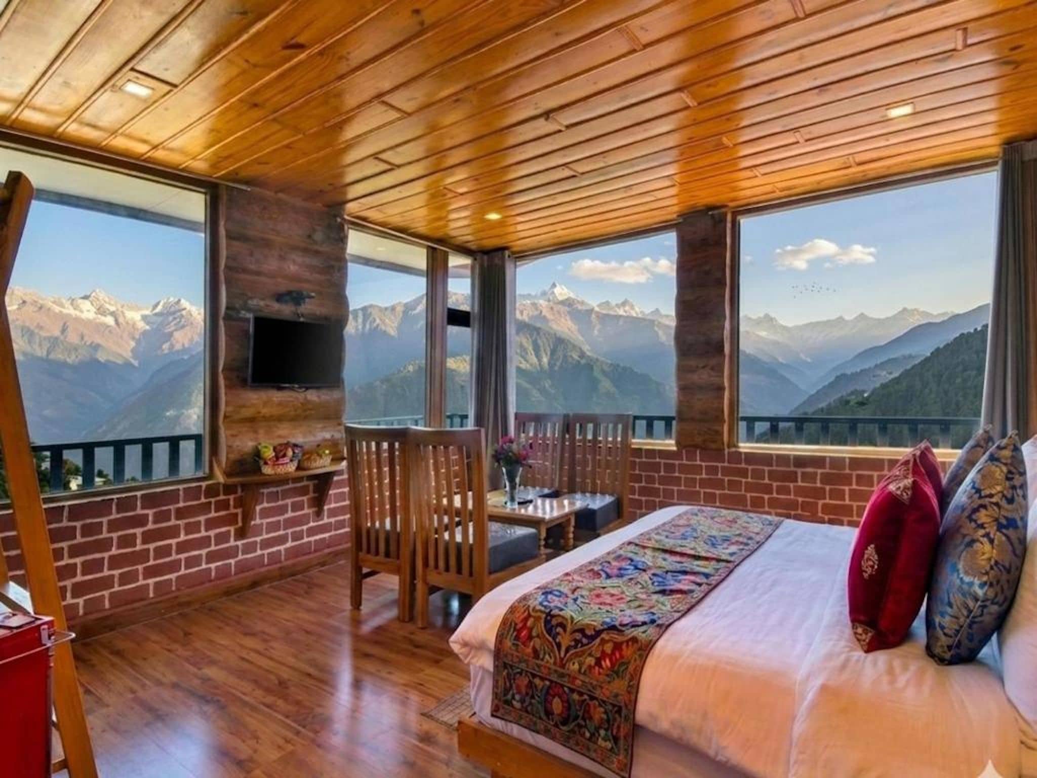 A room with a bed, a TV, a table, and chairs, with mountain views through large windows.