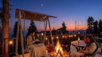 Un grupo de personas sentadas alrededor de un brasero en un patio al aire libre, con una hamaca y luces colgantes.