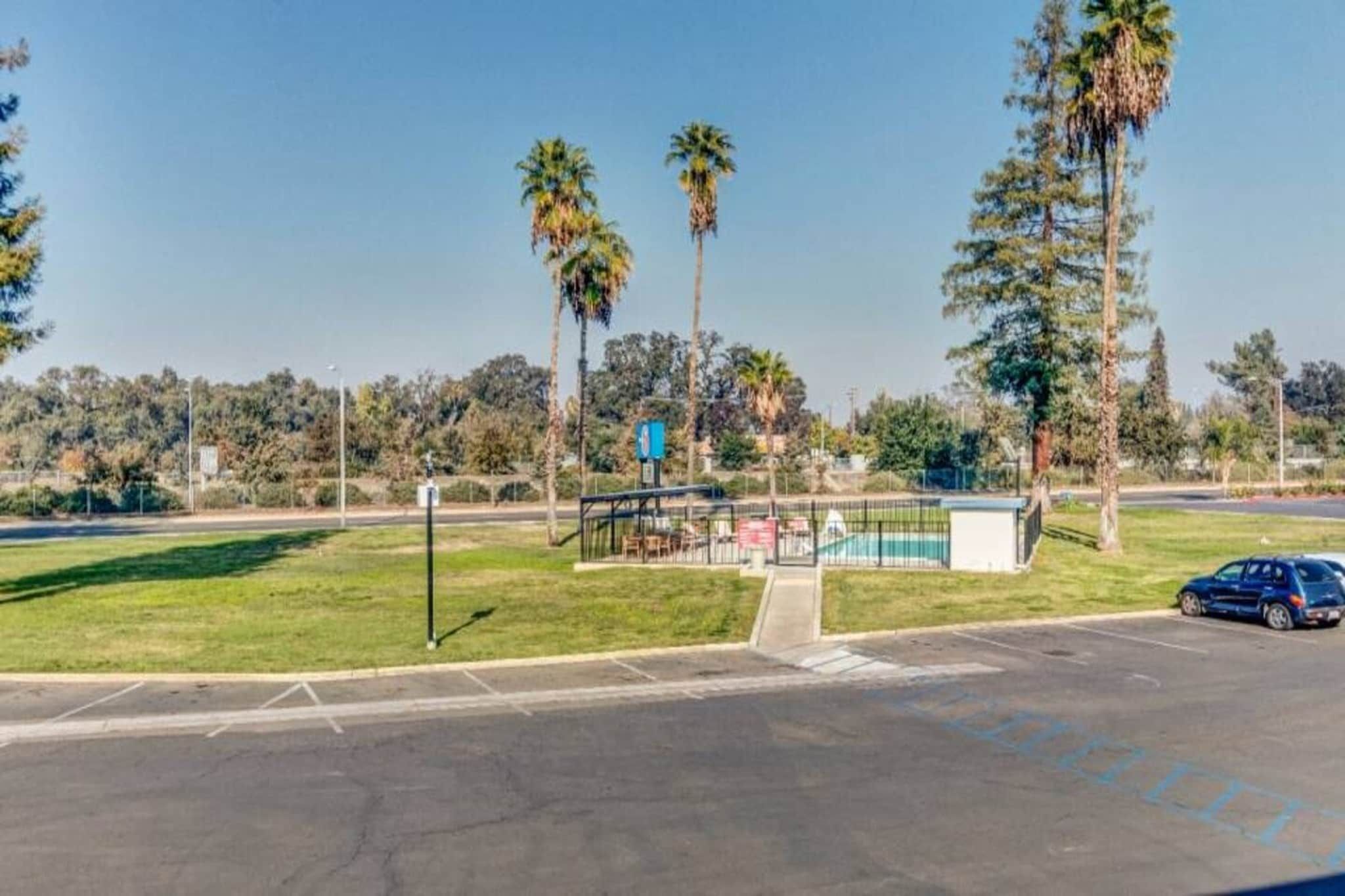 A parking lot with a blue car, a fenced area with a small building, and palm trees in the background.