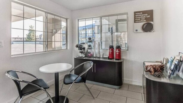 Een lichte, moderne kamer met een witte tafel en twee stoelen, een koffiezetapparaat en een koelkast.