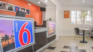 Een receptiegebied van een motel met een groot bord waarop 'motel 6' staat, een balie met een computer en een zithoek met stoelen en een tafel.