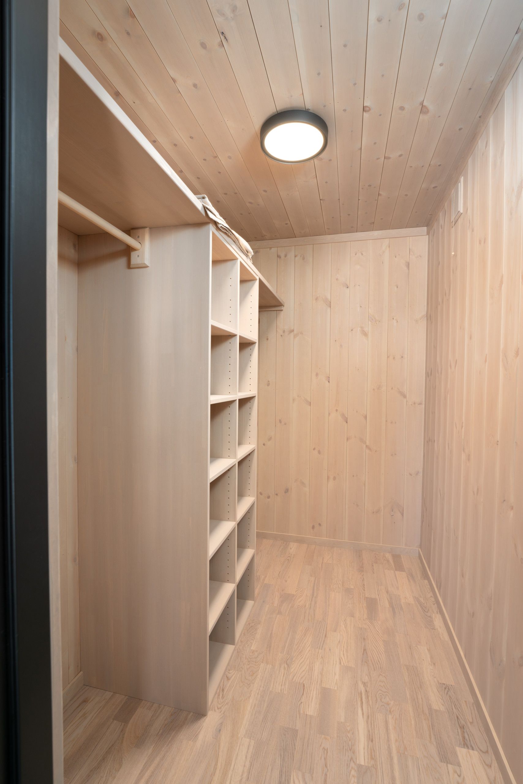 A room with wooden walls and a wooden floor, featuring a built-in closet with multiple shelves and a ceiling light.