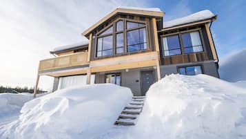 A modern two-story house with large windows and a wooden exterior, surrounded by snow.