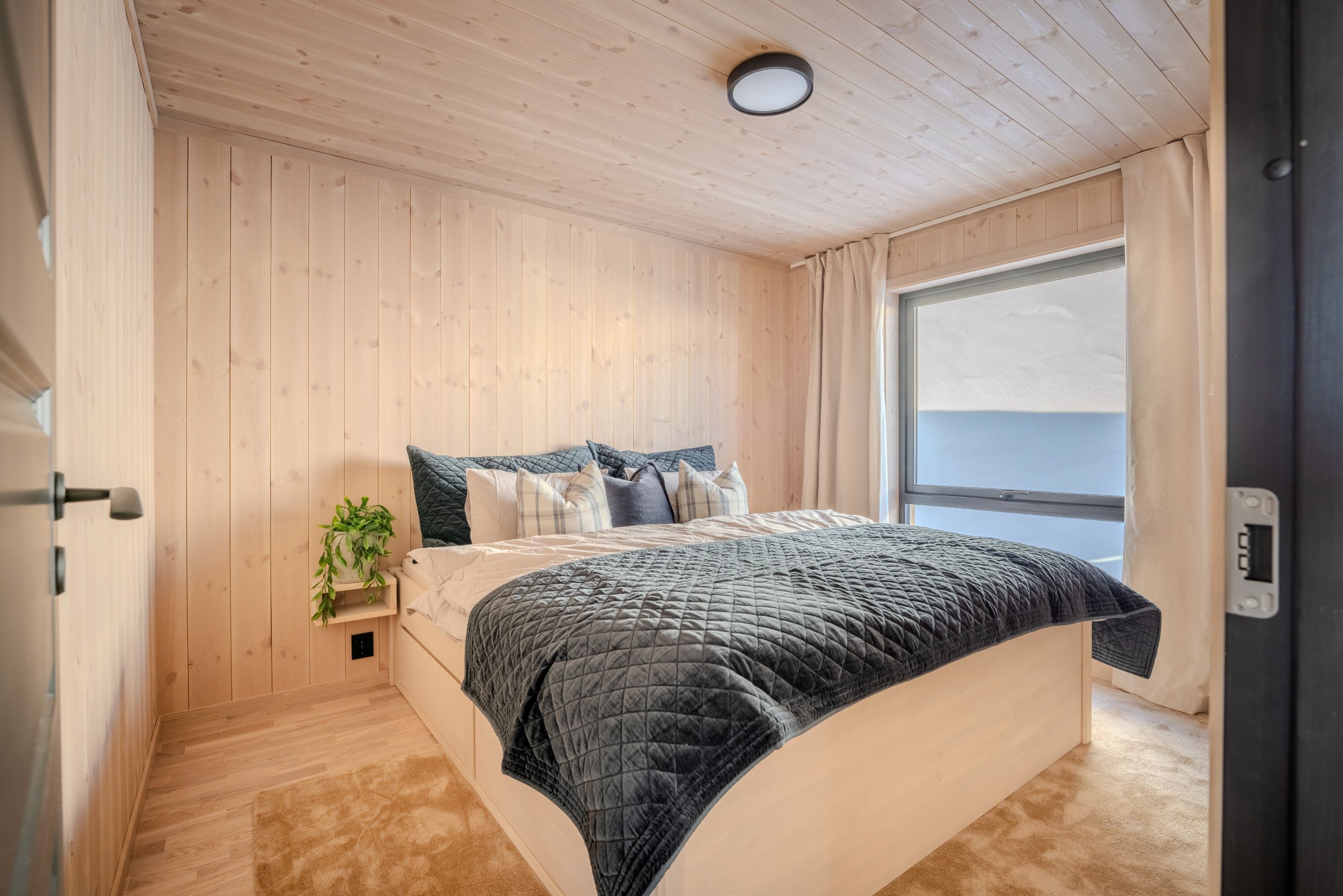 Une chambre à coucher avec un lit, une table de chevet ornée d’une plante, une fenêtre avec des rideaux et un plafond en bois.
