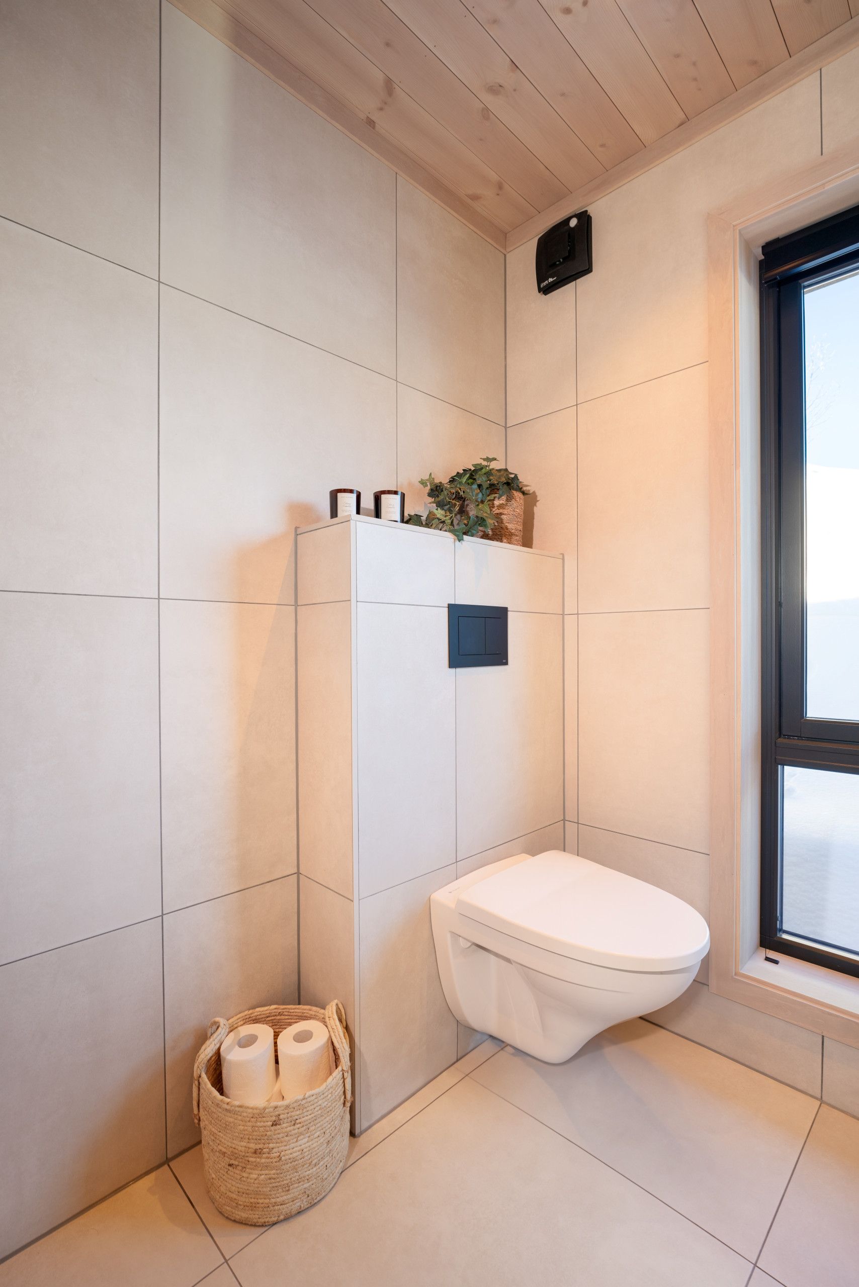 Un baño moderno con inodoro blanco, un ventanal pequeño y un estante en la pared con artículos de aseo.