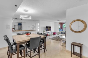 Ein moderner Essbereich mit Holztisch und schwarzen Stühlen, eine Küche mit Kühlschrank und Schränken sowie ein Wohnbereich mit Sofa und Fernseher.