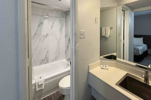 Un baño con una bañera de mármol, inodoro blanco, lavamanos y un espejo que refleja una cama.