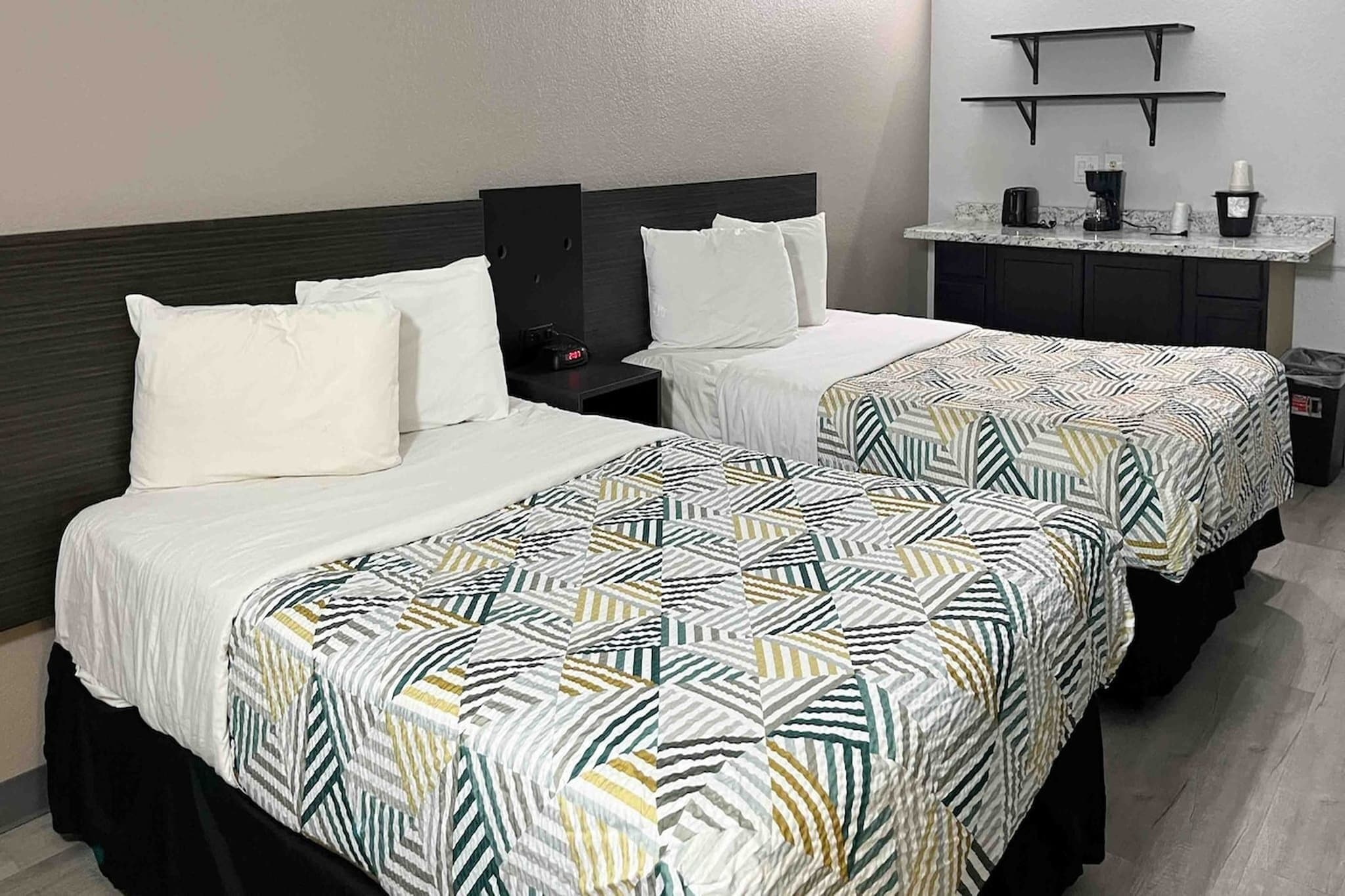 Two single beds with white linens and patterned bedspreads, a nightstand with a clock, and a small kitchenette area with a coffee maker and a trash bin.