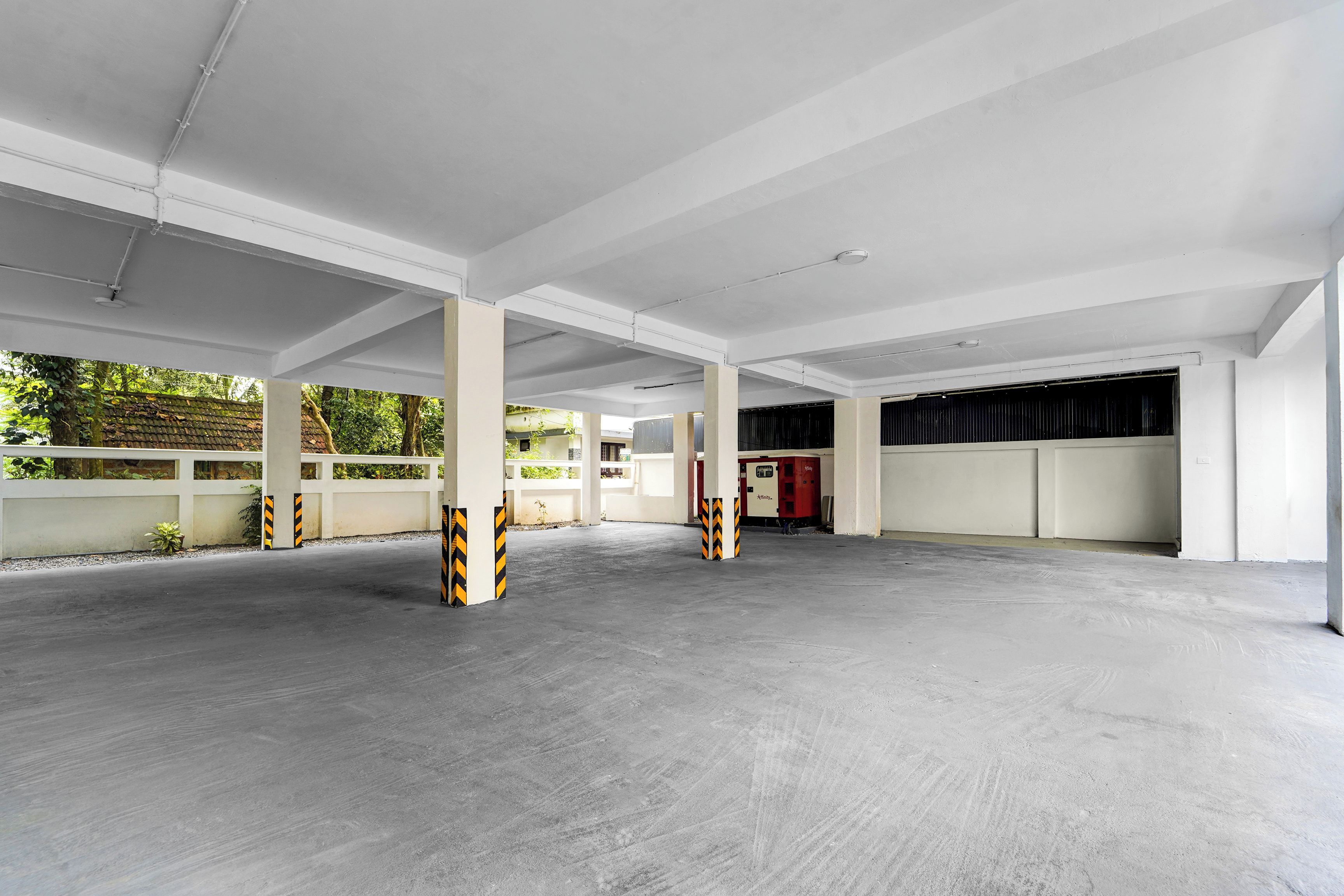 Amplo espaço de estacionamento coberto, com piso de concreto e paredes brancas, com vários pilares listrados em amarelo e preto.