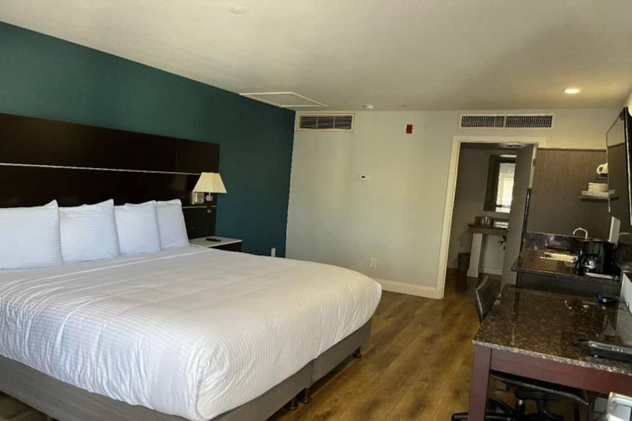 A hotel room with a large bed, a bedside table with a lamp, a bathroom with a mirror, and a kitchenette area.
