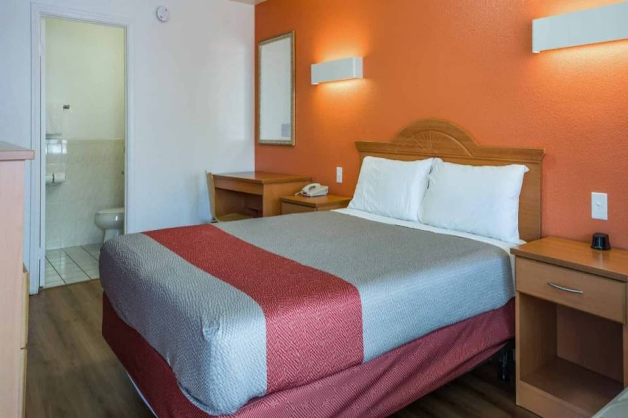 A hotel room with a bed, bedside tables, a nightstand, a mirror, and a bathroom visible in the background.