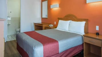 A hotel room with a bed, bedside tables, a nightstand, a mirror, and a bathroom visible in the background.
