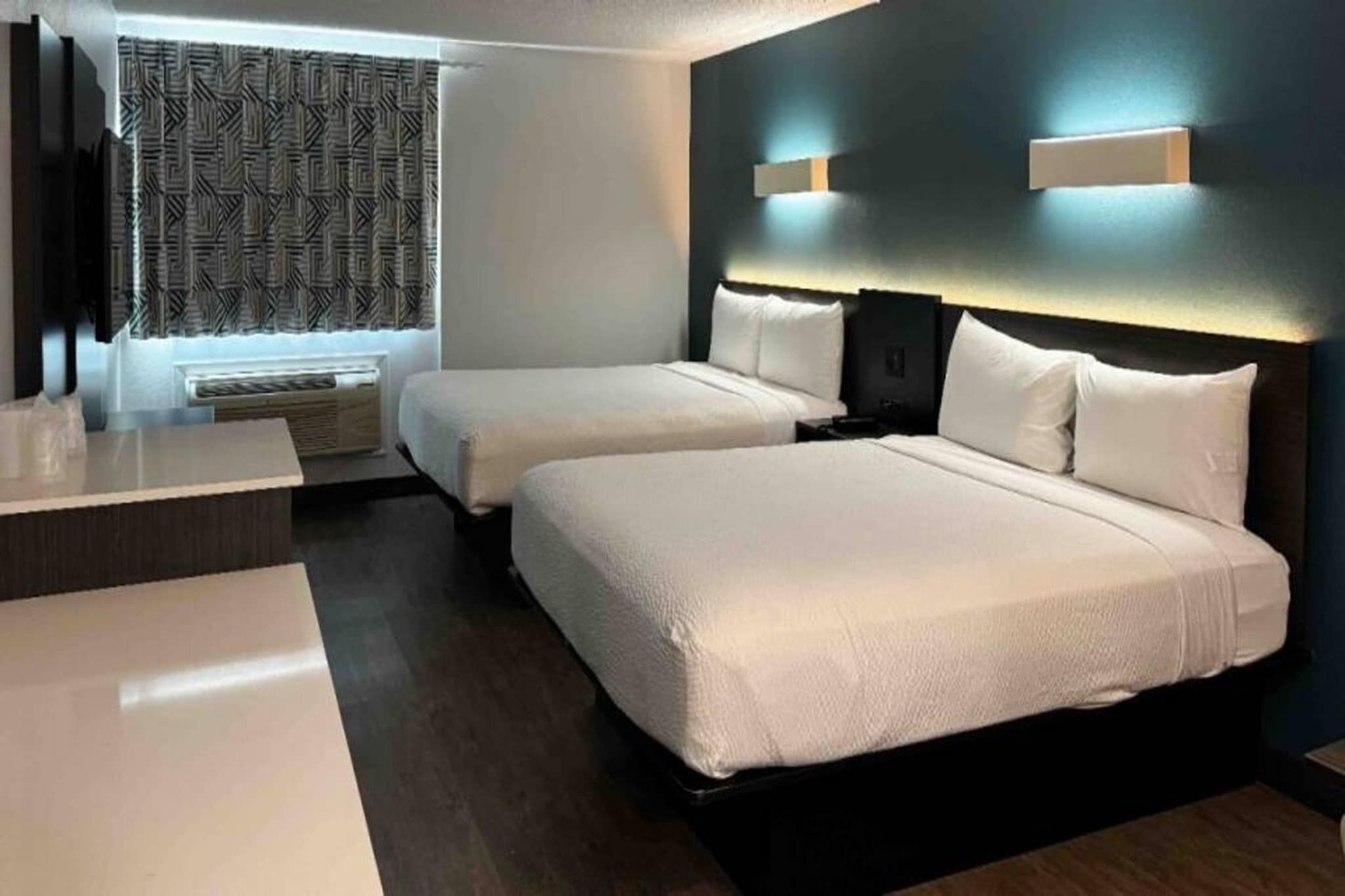 A hotel room with two beds, a desk, and a window with patterned curtains.
